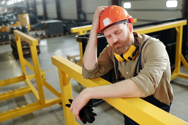 Tired male worker in hard hat leaning on railing on a factory floor.